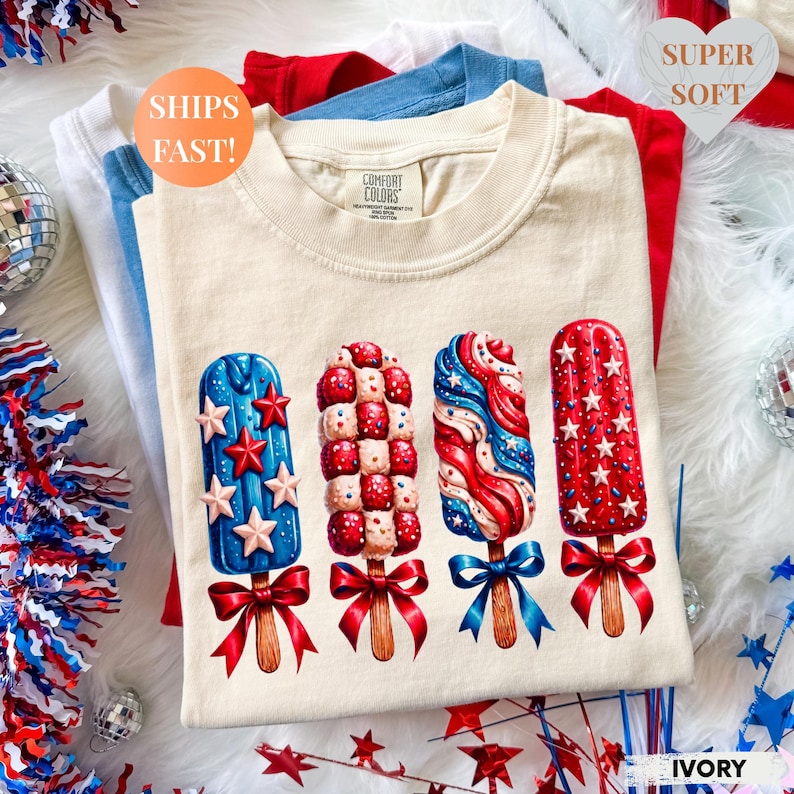 May include: Ivory-colored t-shirt featuring four patriotic popsicle designs in red, white, and blue. The popsicles have star and swirl patterns, with red bows. The shirt is stacked with other shirts in red, white, and blue. The text "SUPER SOFT" is visible.