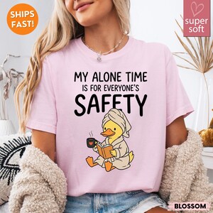K&ouml;nnte beinhalten: Hellrosa T-Shirt mit dem Text "MY ALONE TIME IS FOR EVERYONE'S SAFETY". Das Shirt zeigt eine Cartoon-Ente in einem Bademantel und Handtuch, die ein Buch liest und eine Kaffeetasse h&auml;lt. Das Shirt ist mit "super soft" gekennzeichnet.