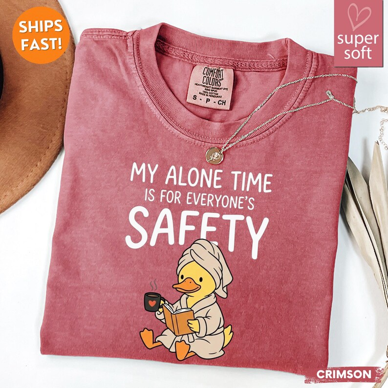 K&ouml;nnte beinhalten: Karmesinrotes T-Shirt mit dem Text "MY ALONE TIME IS FOR EVERYONE'S SAFETY". Das Shirt zeigt eine Cartoon-Ente, die einen Bademantel tr&auml;gt und ein Buch liest, w&auml;hrend sie eine Tasse h&auml;lt. Eine silberne Halskette ist &uuml;ber dem Shirt drapiert.