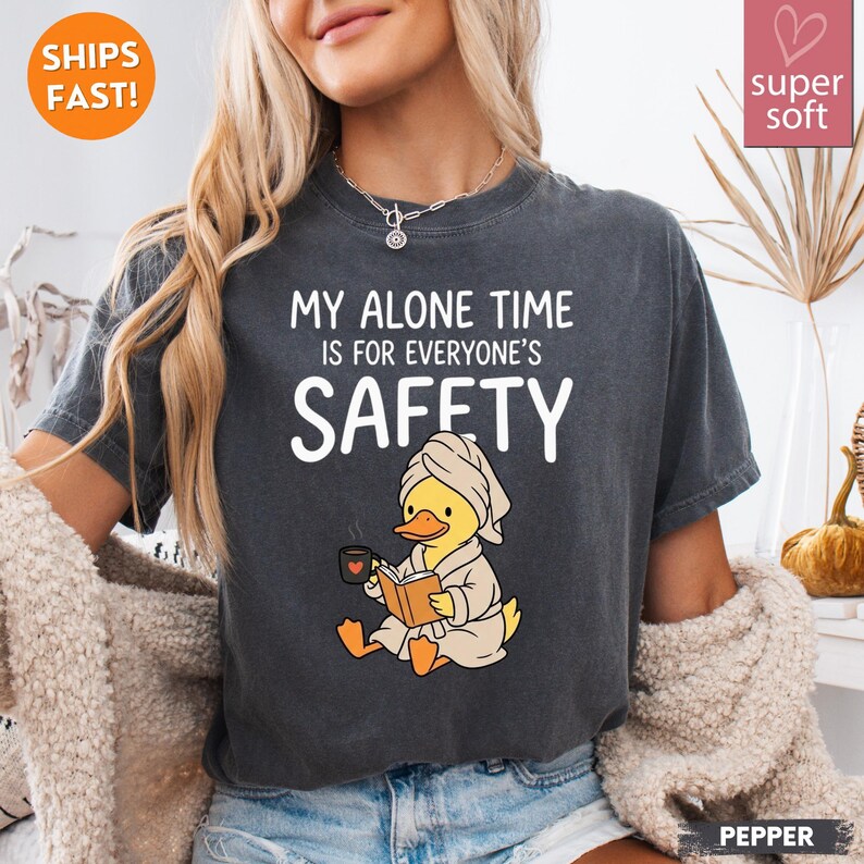 May include: Dark gray t-shirt with the text "MY ALONE TIME IS FOR EVERYONE'S SAFETY." A cartoon duck wearing a robe and towel is reading a book and holding a mug. The shirt has a relaxed fit and is labeled "super soft."