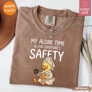 K&ouml;nnte beinhalten: Espressofarbenes T-Shirt mit dem Text "MY ALONE TIME IS FOR EVERYONE'S SAFETY". Das Shirt zeigt eine Cartoon-Ente mit Handtuch und Bademantel, die ein Buch liest und eine Tasse h&auml;lt. Eine silberne Halskette ist &uuml;ber dem Shirt drapiert.