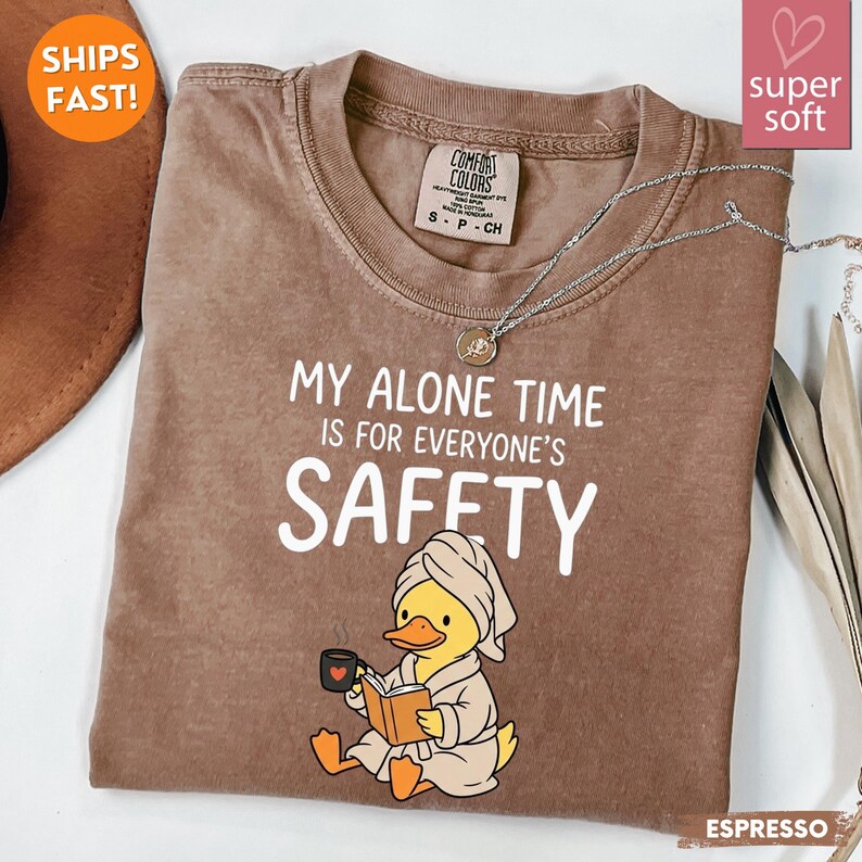 K&ouml;nnte beinhalten: Espressofarbenes T-Shirt mit dem Text "MY ALONE TIME IS FOR EVERYONE'S SAFETY". Das Shirt zeigt eine Cartoon-Ente mit Handtuch und Bademantel, die ein Buch liest und eine Tasse h&auml;lt. Eine silberne Halskette ist &uuml;ber dem Shirt drapiert.