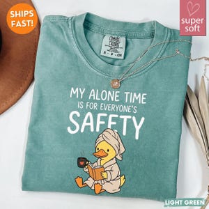 K&ouml;nnte beinhalten: Hellgr&uuml;nes T-Shirt mit dem Text "MY ALONE TIME IS FOR EVERYONE'S SAFETY". Ein Cartoon-Ente im Bademantel liest ein Buch und h&auml;lt eine Tasse. Eine silberne Halskette liegt &uuml;ber dem Shirt.
