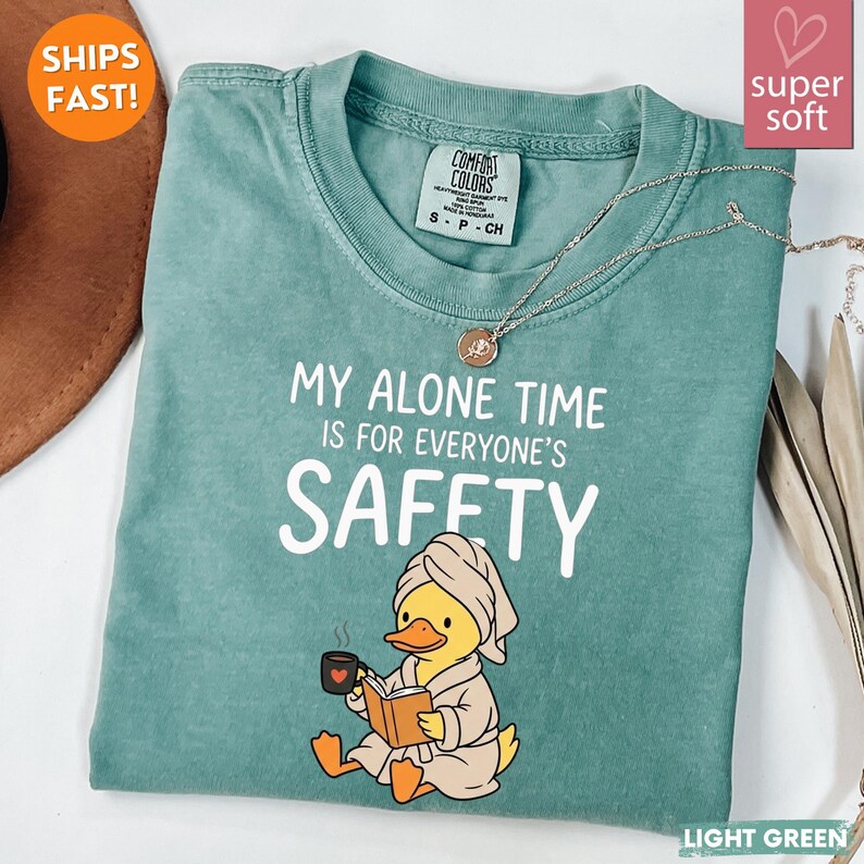 K&ouml;nnte beinhalten: Hellgr&uuml;nes T-Shirt mit dem Text "MY ALONE TIME IS FOR EVERYONE'S SAFETY". Ein Cartoon-Ente im Bademantel liest ein Buch und h&auml;lt eine Tasse. Eine silberne Halskette liegt &uuml;ber dem Shirt.