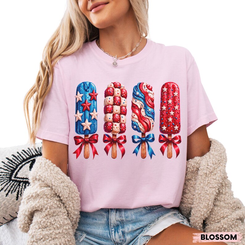 Pu&ograve; includere: T-shirt rosa chiaro con quattro disegni di ghiaccioli rossi, bianchi e blu, con dettagli di stelle e fiocchi. La maglietta ha maniche corte e girocollo. La parola "BLOSSOM" &egrave; stampata in basso a destra.