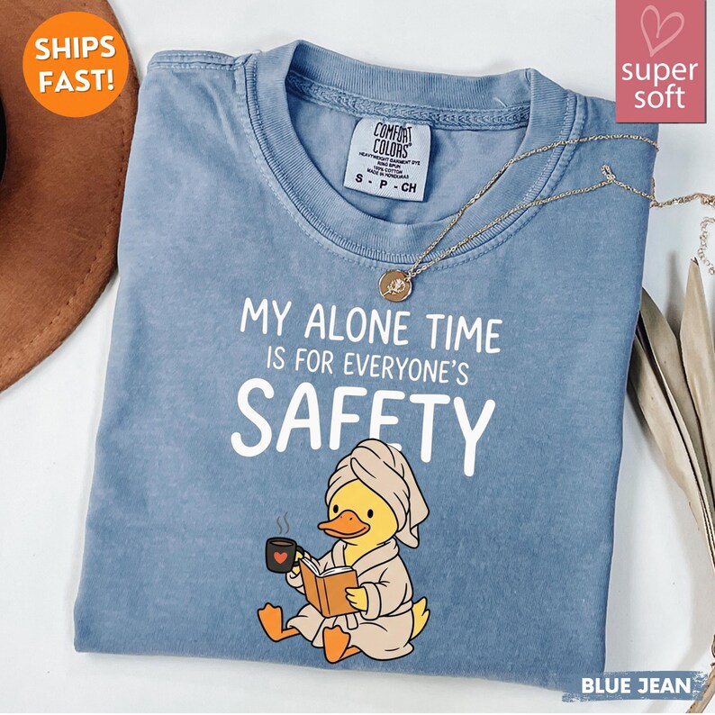 K&ouml;nnte beinhalten: Blaues Jeans-T-Shirt mit dem Text "MY ALONE TIME IS FOR EVERYONE'S SAFETY". Das Shirt zeigt eine Cartoon-Ente mit Handtuch und Bademantel, die ein Buch und eine Tasse h&auml;lt. Eine goldene Halskette ist &uuml;ber dem Shirt drapiert.