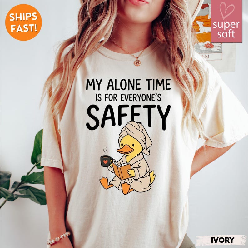 K&ouml;nnte beinhalten: Elfenbeinfarbenes T-Shirt mit dem Text "MY ALONE TIME IS FOR EVERYONE'S SAFETY". Ein Cartoon-Ente mit Handtuch und Bademantel liest ein Buch und h&auml;lt eine Kaffeetasse. Auf dem Shirt stehen auch die Worte "super soft" und "IVORY".
