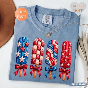Pu&ograve; includere: T-shirt blu jeans Comfort Colors con quattro disegni di ghiaccioli rossi, bianchi e blu. Ogni ghiacciolo ha un bastoncino di legno e un fiocco rosso o blu. La maglietta ha il testo "SUPER SOFT" e "SHIPS FAST!"