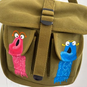 Puede incluir: Un bolso de mensajero de lona color verde oliva con cierre de solapa y hebilla de plástico negro. Dos bolsillos frontales están decorados con ilustraciones pintadas a mano de una criatura rosa a la izquierda y una azul a la derecha, ambas con ojos.