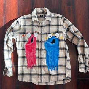 May include: A beige and black plaid button-down shirt with two cartoon monster designs painted on the front. One monster is red, and the other is blue. The shirt has long sleeves and a collared neckline.