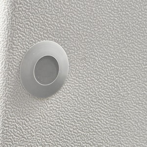 Może przedstawiać: Okrągła, metaliczna srebrna oprawa oświetleniowa wpuszczana w białą, teksturowaną, ziarnistą powierzchnię. Lampa posiada gładki, płaski pierścień zewnętrzny otaczający lekko wpuszczoną, okrągłą soczewkę środkową, tworzącą zlicowany wygląd.