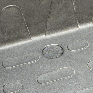 Peut inclure: Un tapis de sol automobile noir présentant une surface texturée avec des lignes horizontales en relief et une série d'indentations ovales répétitives. Un trou de fixation circulaire avec un insert gris foncé texturé est visible au centre.