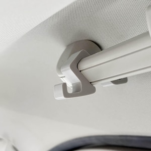 Puede incluir: Primer plano de un accesorio interior de coche blanco. La imagen muestra un gancho de plástico blanco fijado a una barra blanca, montado en un techo blanco texturizado. El gancho está diseñado para sujetar objetos.