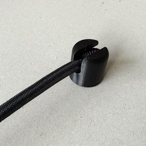 Pode incluir: Um clipe cilíndrico preto para cordão com um entalhe superior, segurando um cordão elástico preto canelado. O clipe apresenta uma base circular e uma ranhura vertical projetada para prender o cordão no lugar, mantendo uma tensão constante.