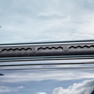 Può includere: Primo piano di un componente nero e scanalato con diversi elementi di fissaggio rotondi grigio scuro. Il componente è posizionato su uno sfondo di cielo nuvoloso, con riflessi visibili su una superficie lucida sottostante.