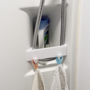 Può includere: Un organizer per doccia bianco con un tubo doccia cromato e una bottiglia di shampoo. Due piccoli asciugamani sono appesi all'organizer, uno con un anello arancione e l'altro con un anello blu. L'organizer è montato su una parete doccia bianca.