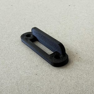 Op de afbeelding: Een zwarte, 3D-geprinte beugel met een rechthoekige opening en twee ronde gaten. De beugel heeft een getextureerd oppervlak en een scharnierend gedeelte. Het object is ontworpen voor functioneel gebruik.