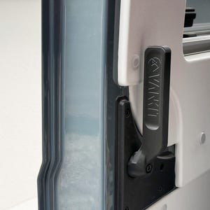 Puede incluir: Un mango vertical negro con el texto VANKIT en relieve montado sobre un panel blanco. El mango está colocado junto a un componente de marco vertical gris oscuro translúcido. El conjunto cuenta con un mecanismo de bisagra con tornillos.