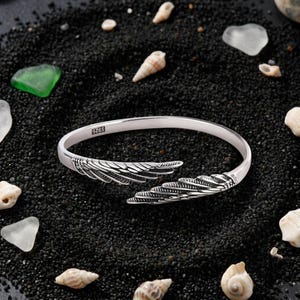 Könnte beinhalten: Ein silberfarbenes, offenes Armband mit detaillierten Flügelmotiven an beiden Enden. Die Metalloberfläche zeigt gravierte Federstrukturen und einen deutlichen S925-Stempel auf der Innenseite, der auf Sterlingsilber hinweist.