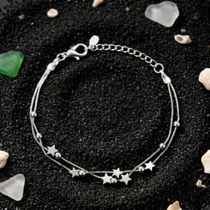 May include: A silver anklet with a double-strand design. The anklet features small star charms and tiny ball accents. The jewellery is displayed on a black surface with small shells and sea glass.