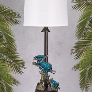 May include: A decorative table lamp featuring a bronze-colored base and stem, topped with a white lampshade. The lamp's base is adorned with three teal-colored turtle figurines, adding a coastal theme. The lamp stands approximately 28 inches tall.