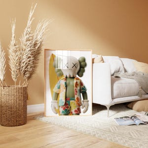 May include: Framed artwork featuring a cartoon figure with a skull-like head, wearing a Hawaiian shirt and shorts, and holding a surfboard. The figure is in front of a yellow surfboard. The artwork is in a light wood frame.