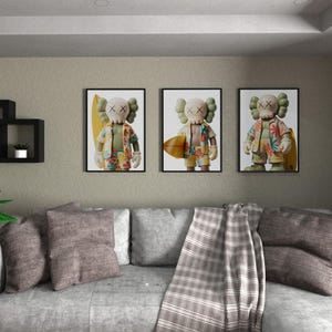 May include: Three framed art prints featuring a cartoon figure with X-shaped eyes and a floral shirt, holding a surfboard. The prints are displayed above a gray sofa with throw pillows and a plaid blanket. A decorative wall shelf and a potted plant are also visible.