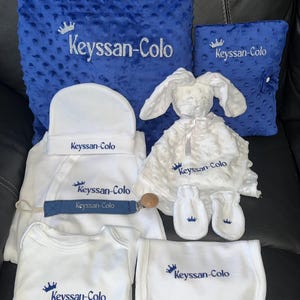 Peut inclure: Un ensemble d'articles pour bébé comprenant une couverture bleue à pois, un carnet bleu, un bonnet blanc, un body blanc, un bavoir blanc, des moufles blanches, un doudou lapin blanc et une attache-sucette bleue, brodés Keyssan-Colo.