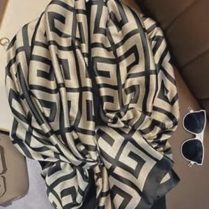 Könnte beinhalten: Beige-schwarzer Schal mit geometrischem Muster. Der Schal ist drapiert und gefaltet, wodurch das komplizierte Muster zur Geltung kommt. Eine Sonnenbrille mit weißem Rahmen und schwarzen Gläsern liegt neben dem Schal.