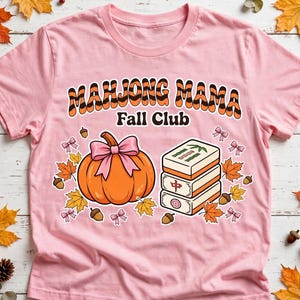 May include: A light pink t-shirt featuring the text "MAHJONG MAMA Fall Club" in orange and black striped lettering. The graphic includes an orange pumpkin with a pink bow, a stack of three mahjong tiles, scattered autumn leaves, and acorns.