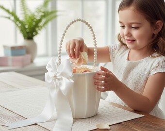 Op de afbeelding: Een witte bloemenmeisjesmand met een parelmoeren handvat en een grote satijnen strik. De mand is gevuld met rozenblaadjes. De mand staat op een houten tafel.