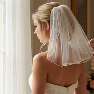May include: A sheer, white bridal veil with delicate pearl accents. The veil is held in place, showcasing its soft, flowing fabric and subtle embellishments. The bride is wearing a strapless white dress.