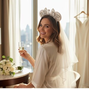 May include: A bride-to-be wearing a white satin robe and a "BRIDE" headband with sparkling embellishments. She holds a champagne flute, smiling at the camera. A bouquet of white roses and a wedding dress are in the background.