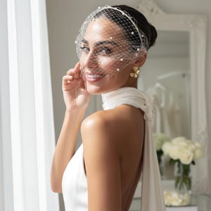 Peut inclure: Une mariée porte une robe blanche et un voile cage à oiseaux orné de perles. Le voile est fixé avec un bandeau. La mariée porte également des boucles d'oreilles dorées en forme de fleur. La robe a un col en licou.