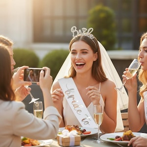 Puede incluir: Una futura novia celebra con amigos al aire libre. Lleva una banda blanca que dice "bride to be" y una tiara brillante que dice "Bride". La novia se ríe y levanta las manos. Otras personas están tomando fotos y bebiendo champán.