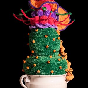 Può includere: Una scultura di cactus stravagante, realizzata a mano, in filato verde, arancione, viola e rosso. Il cactus è adornato con spilli arancioni e si trova in un vaso di ceramica bianca con manici. Lo sfondo è nero.