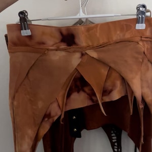 May include: A distinctive brown skirt with an irregular, layered design. The skirt has a tie-dye effect and a black panel with gold buttons. The skirt is suspended from a white hanger.