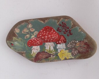 Pode incluir: Pedra pintada à mão com três cogumelos de chapéu vermelho com pintas brancas, sobre um fundo de folhas verdes e folhagem de outono. Uma pequena joaninha é visível no lado esquerdo. A pedra tem uma borda marrom.