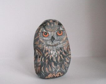 Pode incluir: Estatueta de coruja de pedra pintada à mão. A coruja é pintada em tons de cinza, marrom e laranja, com padrões de penas detalhados e olhos laranja brilhantes. A pedra tem formato oval e fica em pé.