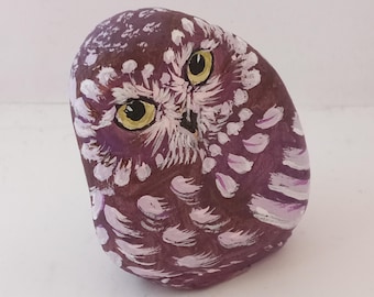 Pode incluir: Uma pedra pintada à mão em forma de coruja. A coruja é pintada em tons de roxo e branco, com olhos amarelos. A pedra é lisa e arredondada, e os detalhes da coruja são bem definidos.