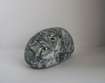 Pode incluir: Uma pedra pintada à mão em cinza e branco com uma representação realista do rosto de um gato. O gato tem olhos verdes e um nariz rosa.