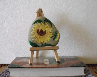 Pode incluir: Uma pedra pintada com um desenho de girassol em um pequeno cavalete de madeira. A pedra é cinza com folhas verdes e um girassol amarelo com um centro marrom. O cavalete é de madeira marrom.