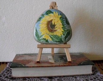 Pode incluir: Uma pedra pintada com um desenho de girassol em um pequeno cavalete de madeira. A pedra é pintada com um girassol realista com pétalas amarelas e um centro marrom. O fundo é de cor azul claro. O cavalete é feito de madeira e tem um acabamento natural.