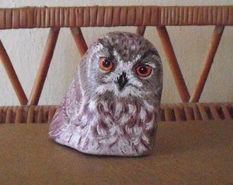 Pode incluir: Estatueta de coruja de pedra pintada à mão. A coruja tem olhos grandes alaranjados, um bico preto e um padrão de penas detalhado em tons de castanho, branco e roxo. A coruja está sobre uma superfície castanha.