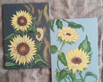 Pode incluir: Duas pequenas pinturas de girassóis. Uma pintura tem um fundo cinza escuro com girassóis amarelos e castanhos e folhas verdes. A outra pintura tem um fundo azul claro com girassóis amarelos e folhas verdes.