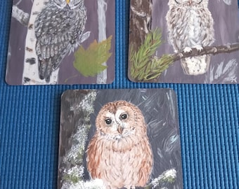 Pode incluir: Três bases quadradas de madeira com desenhos de corujas pintados à mão. Cada base apresenta uma espécie diferente de coruja empoleirada num galho, contra um fundo cinza escuro com detalhes de árvores e folhas. As corujas são retratadas em tons de cinza, castanho e branco.