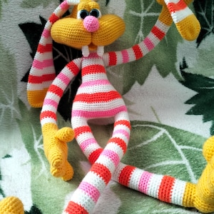 May include: A handmade, crocheted bunny toy with a yellow head, ears, and feet. The bunny's body and limbs are striped with white, pink, and orange. It has large, blue eyes and a pink nose. The toy is designed to be soft and cuddly.