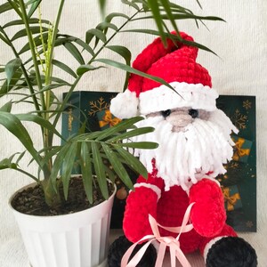 May include: A handmade plush Santa Claus doll with a red hat and suit, white beard, and black boots. The doll is tied with a pink ribbon. A potted plant is in the background.
