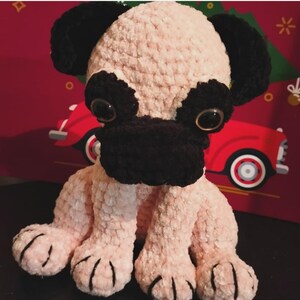 May include: A handmade crochet pug dog plushie. The dog has a light pink body, black ears, muzzle, and paws. It has large, round, clear eyes. The plushie is sitting upright. The background is a red and green holiday scene.