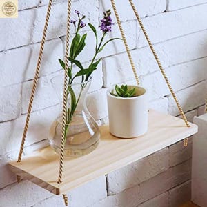 Könnte beinhalten: Hängendes Holzregal mit natürlichen Seilhaltern. Eine klare Glasvase mit lila Blumen und ein kleiner weißer Topf mit einer Sukkulente stehen auf dem Regal. Das Regal befindet sich vor einer weißen Backsteinmauer.